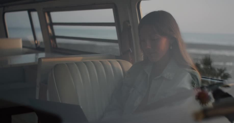 A woman sitting inside a van typing on a laptop overlooking the beach