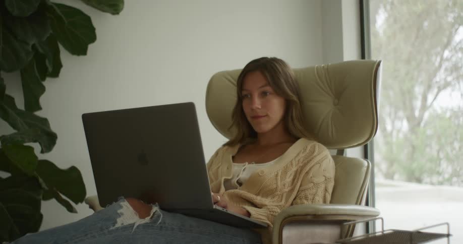 Young woman wearing a sweater types on a laptop while sitting in a chair with a headrest