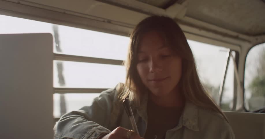A woman takes notes while studying and using her laptop in her van