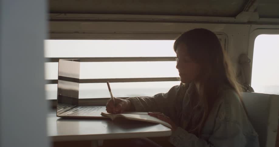 A young woman writes in a notebook while referencing a laptop as she sits inside a recreational vehicle