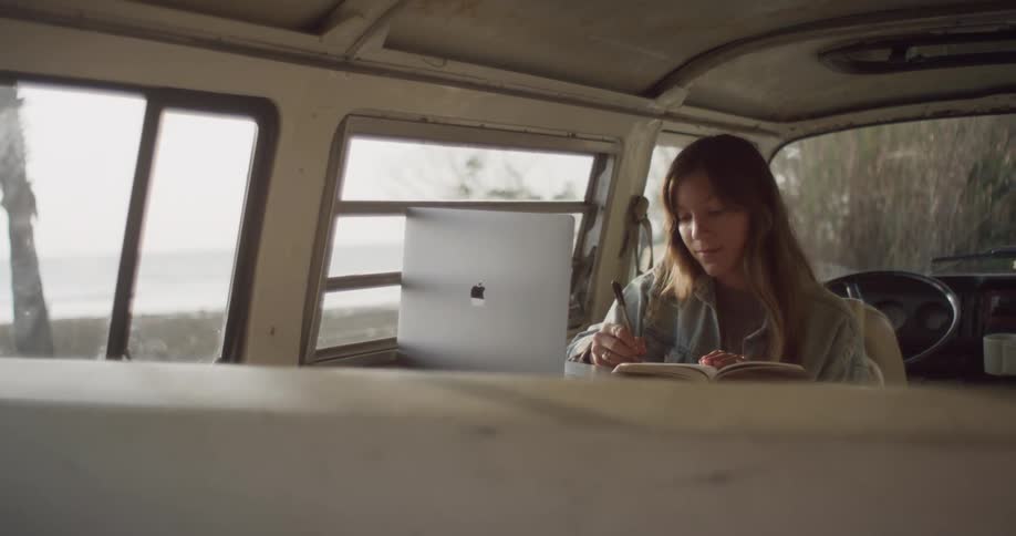 Young woman sitting inside a van parked near the beach takes notes on a notepad from a laptop