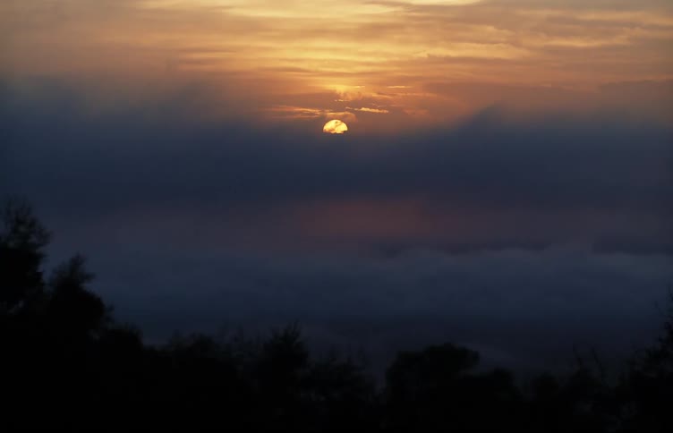 The sun hangs low in the sky and is partially obscured by a thick layer of clouds