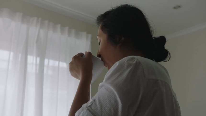 A woman stands in front of a window reflecting on her thoughts as she drinks tea