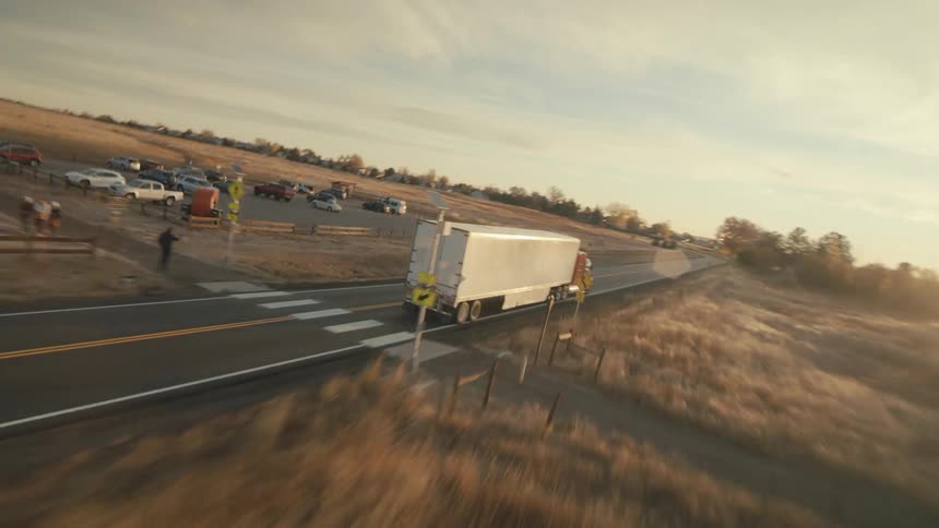 A semi trailer truck travels on a rural road heading toward water with sun glaring