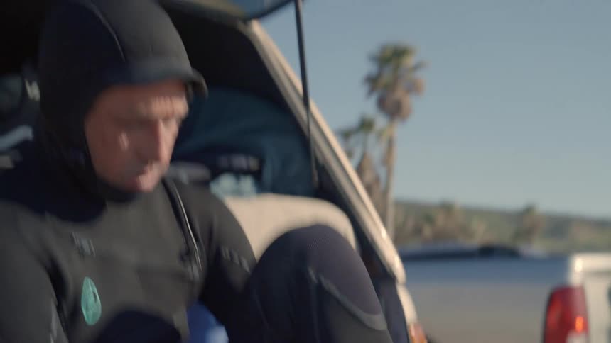 A man wearing a wet suit sitting on his tailgate puts on his booties