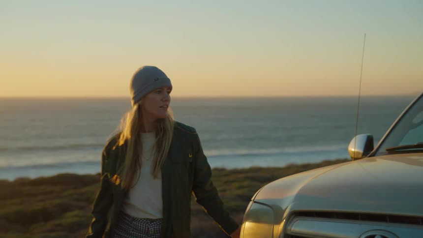 A woman gets out of a truck parked at the ocean and walks skips around it