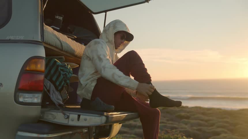 A man sits in the back of his pickup truck by the ocean and puts on his shoes