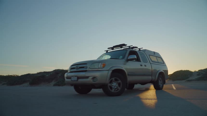 Man drives vehicle out of parking space as sun sits just above horizon shining over nearby vegetation topped sandy mounds