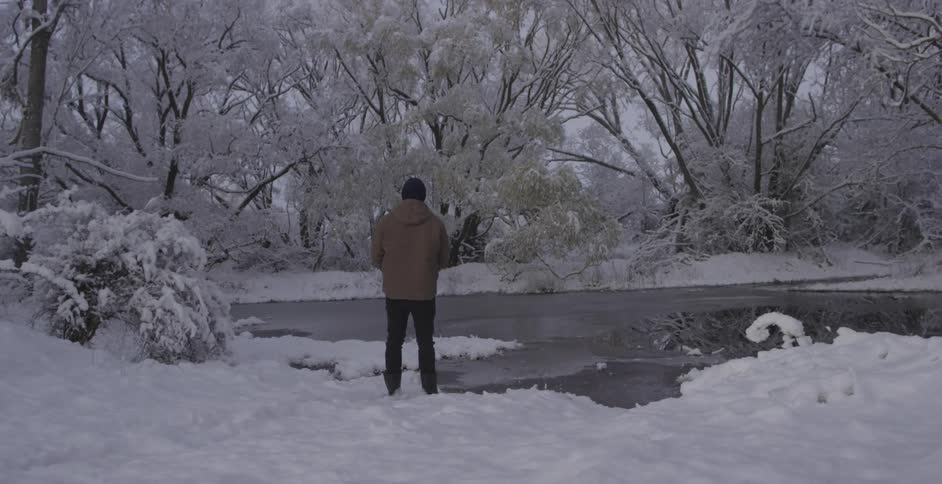 A man stands by a partially ice-covered stream in a snowy wilderness