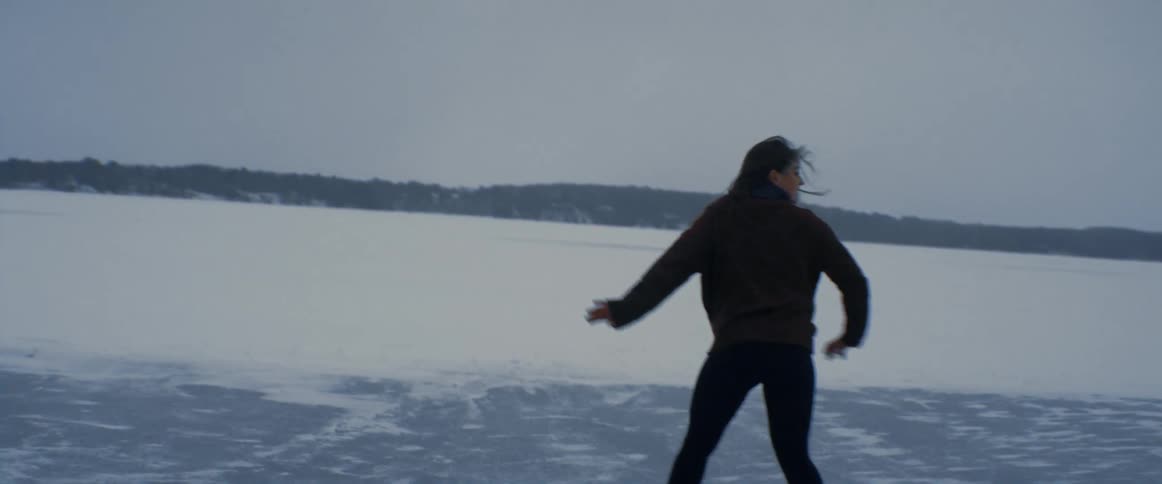 A woman ice skates on a frozen lake surrounded by snow covered fields