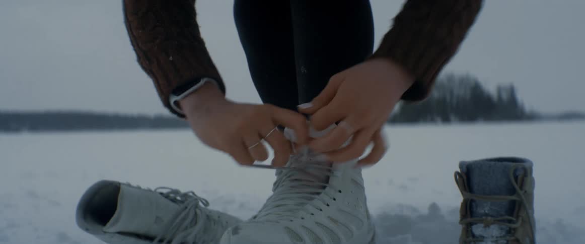 A woman laces up her ice skate bent over standing in the snow