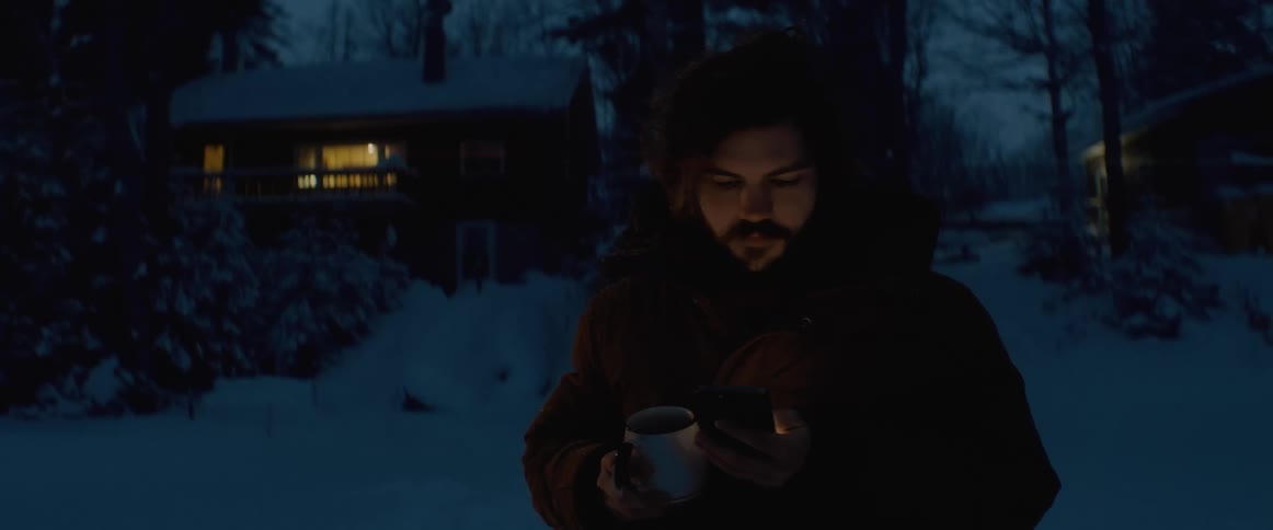 A man scrolls through his phone and drinks coffee while standing outside his house in the snow