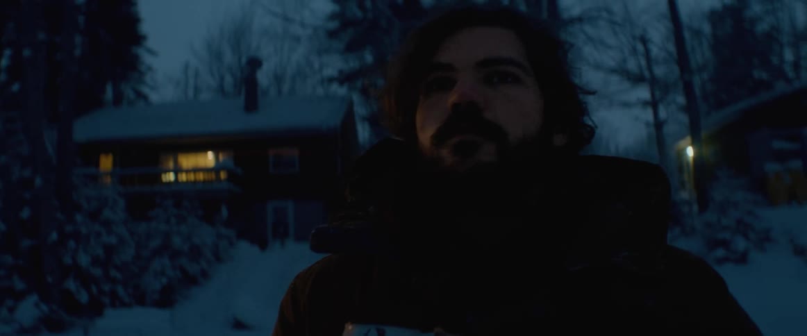 Young bearded man drinking from a cup in front of a house in the snow covered forest at evening