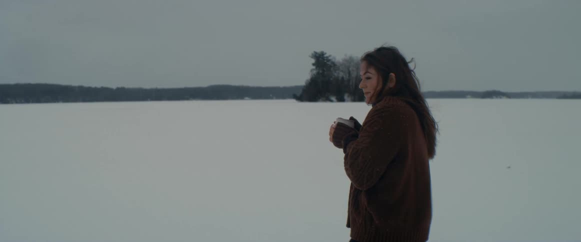 A woman stands outside on a snow covered frozen lake holding a mug while the wind blows