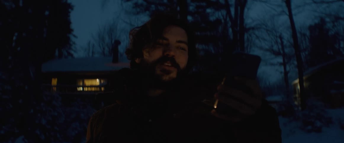 A man pauses talking to sip from a mug while standing outside in the snow with a cabin behind