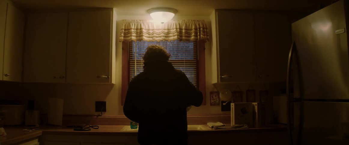 Man with wavy hair and black hoodie pours hot water into cup in dim lit kitchen