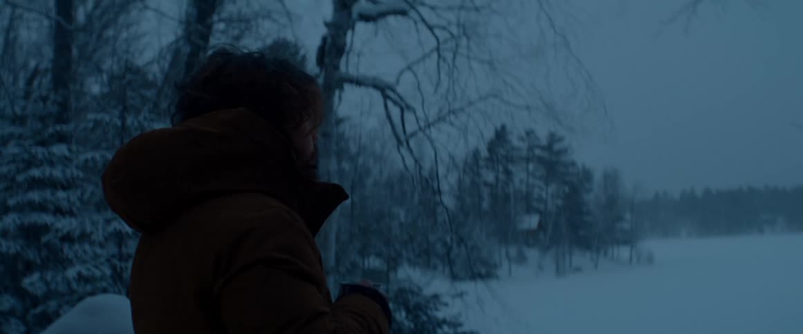 A man stands looking out at the snow and gray sky drinking from a mug