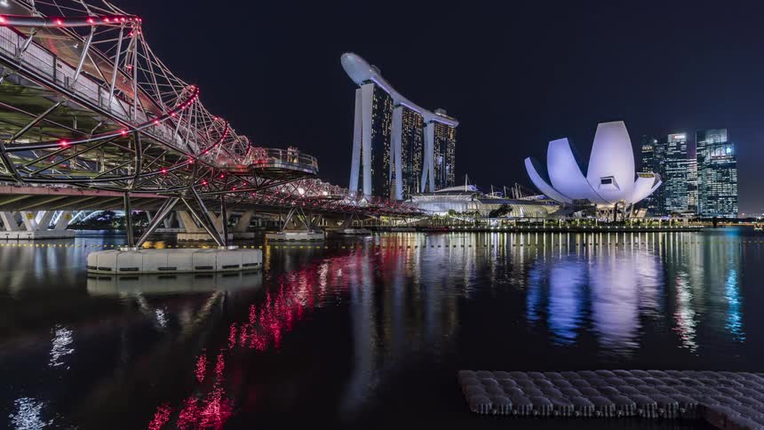 Pedestrians stand near rail on bridge overlooking reflective water near artistically architecture buildings as boats move