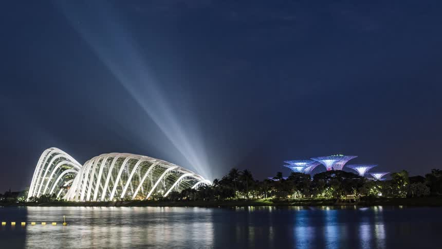 Boats move across water reflecting light from brightly lit buildings with artistic architecture as spotlights shine into the sky