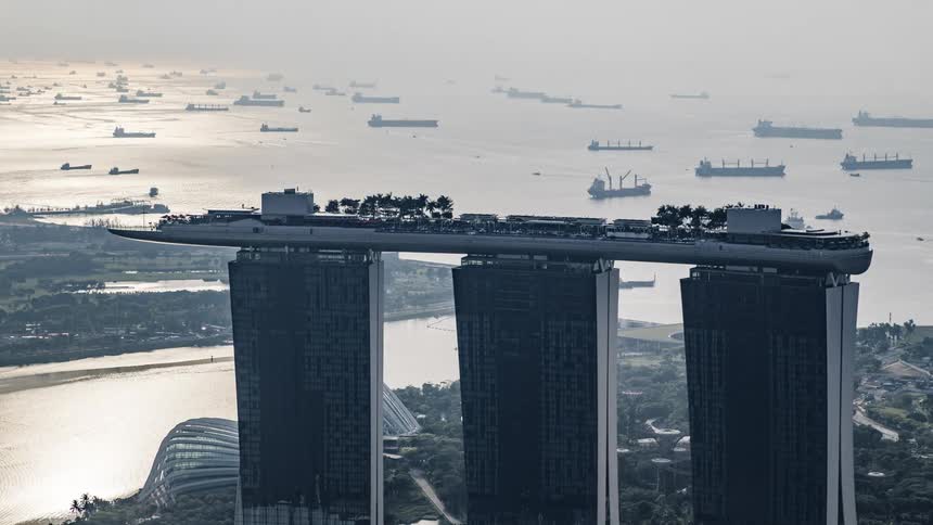 Boats sail through the sea past a set of three tall skyscrapers with a boat on top