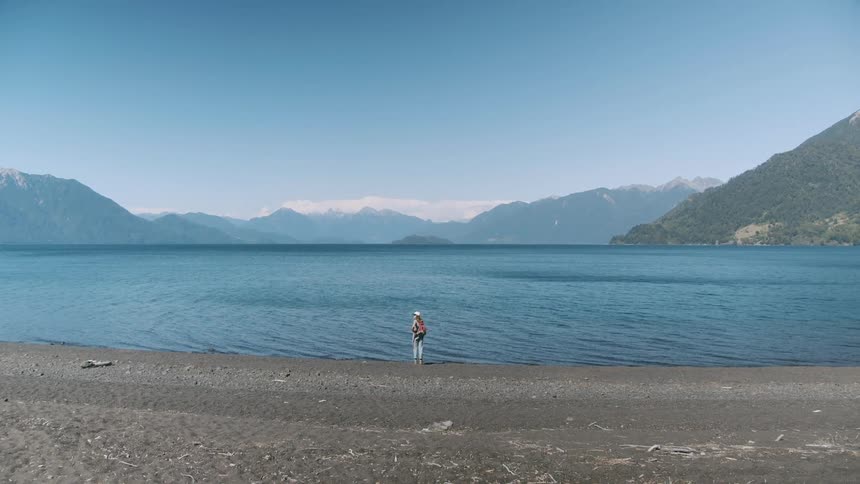 A woman on the beach walks towards the water and bends down and wets her hands and rubs her neck