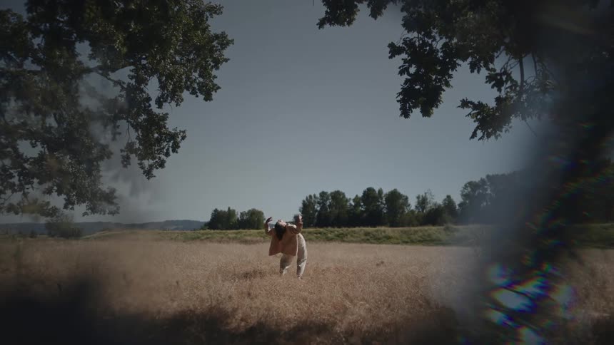 Woman in a jacket and pants stands in a field and bends her body backwards as she spreads her arms