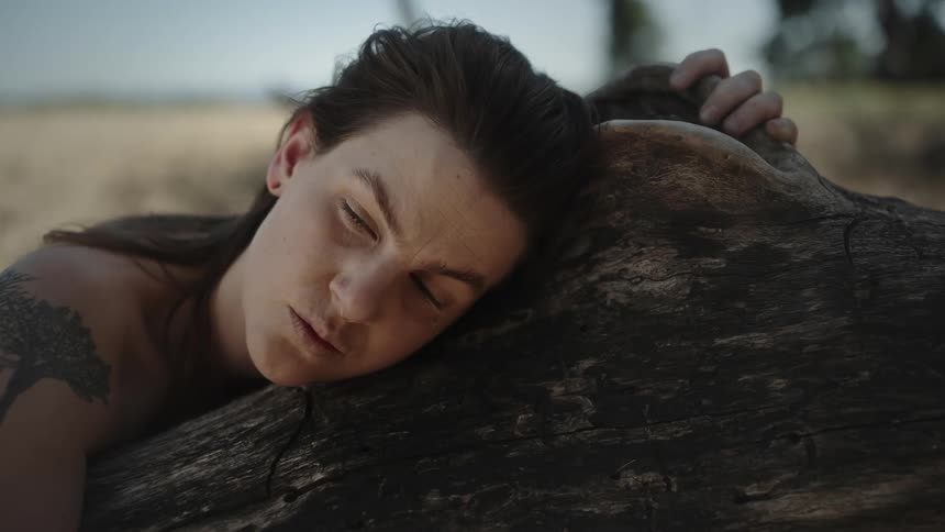 Woman with tattoo and dark hair leans on tree trunk staring into space