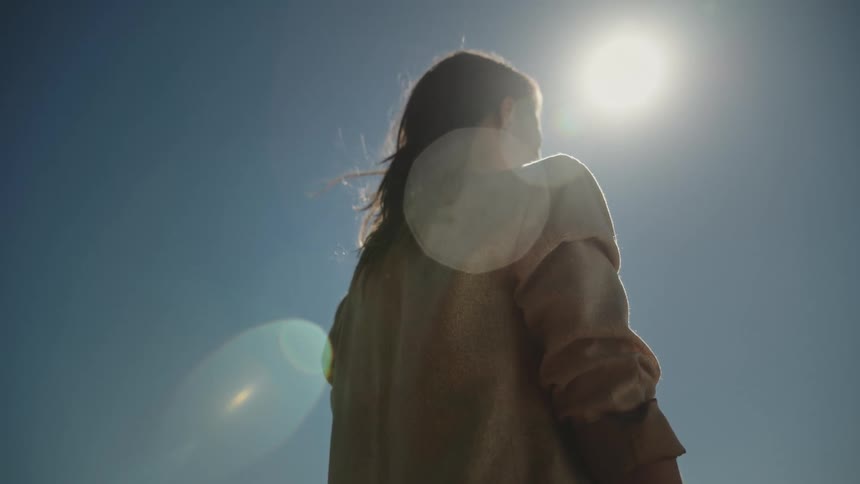 Woman with brown jacket and wavy hair stands under the sun and blue sky