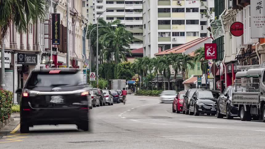 Man stands on back of parked truck as traffic including a double decker bus passes on street bordered by storefronts