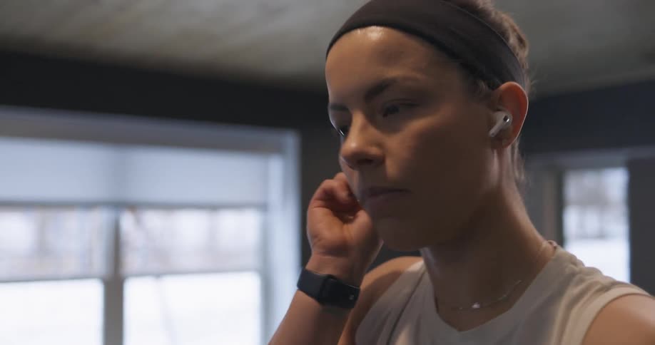 Woman with her hair in a bun and wearing a headband inserts white earbuds into her ears