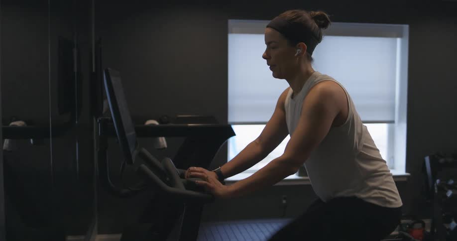 Woman covered in sweat wearing sports clothes working out over a static bicycle at home near a window