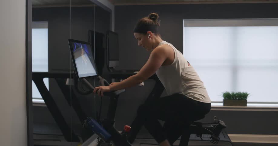 Woman with black headband gray sports shirt and black leggings works out on exercise bike in the gym