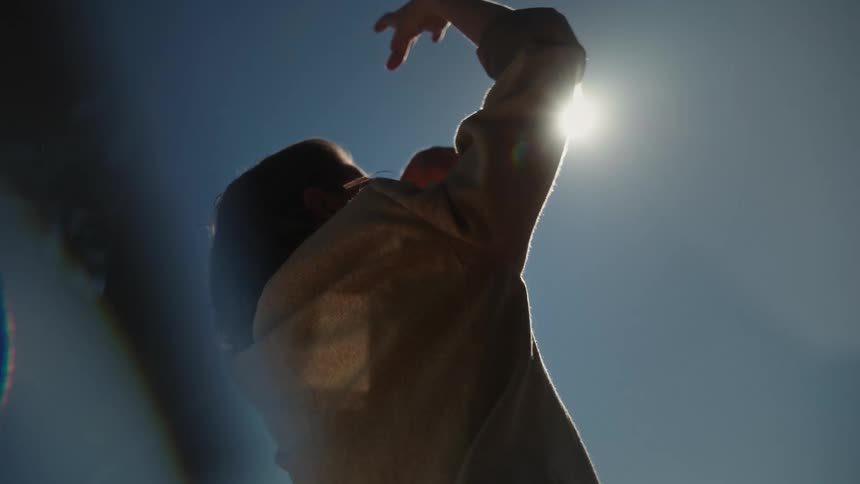 Woman with wavy hair and brown jacket dances under the sun and blue sky
