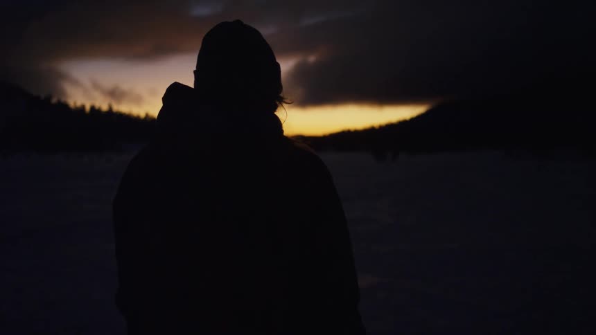Silhouette of a man standing on a frozen field covered in snow watching the sunset