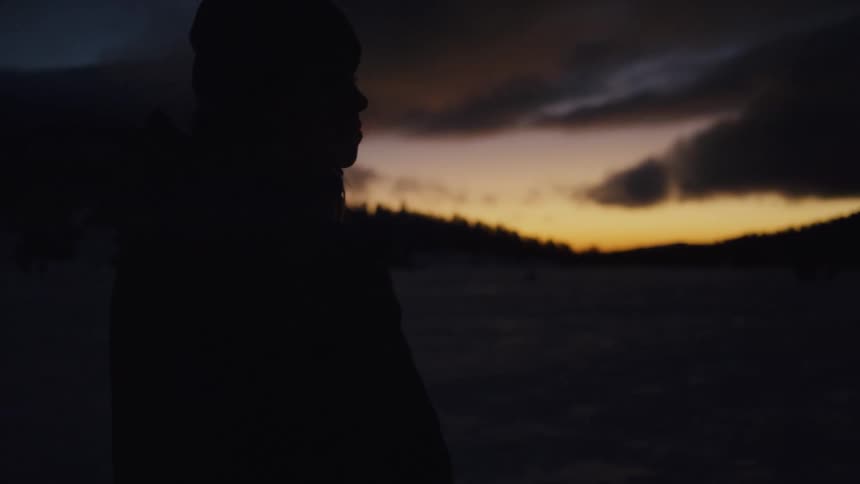 Woman with wooly hat and coat stands on snowy hill and is silhouetted against the sunset