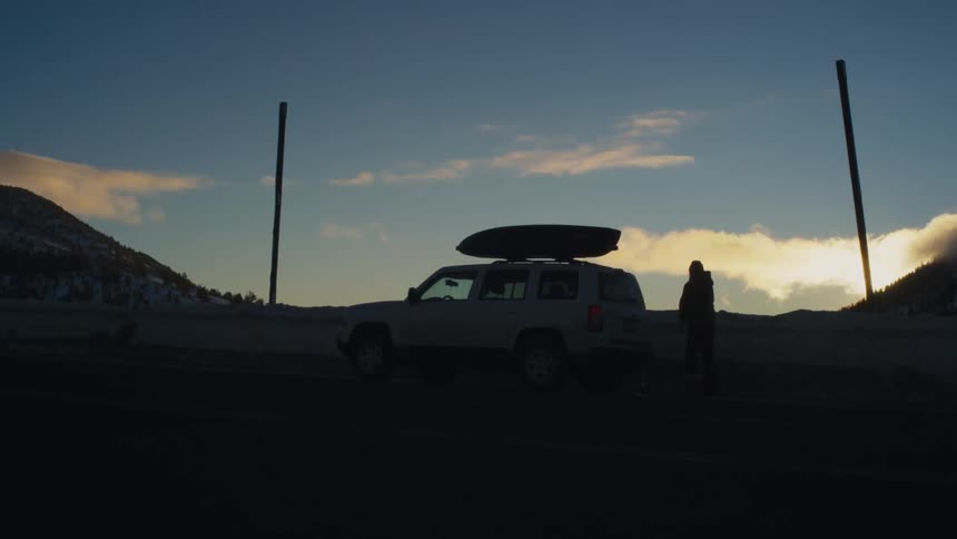 Man closing car boot is silhouetted against the sky at dusk