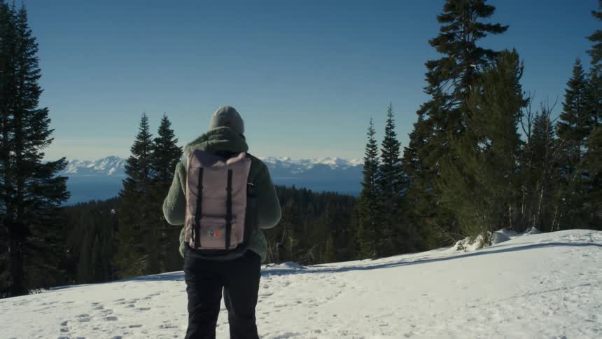 Woman with snow shoes green woolly hat coat black pants and beige backpack walks down snowy hill overlooking forest and distant snowy mountain range