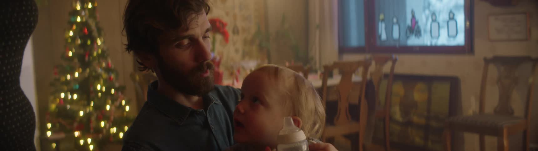 Toddler drinks from a sippy cup while sitting on his fathers lap then hands the cup to an outstretched hand