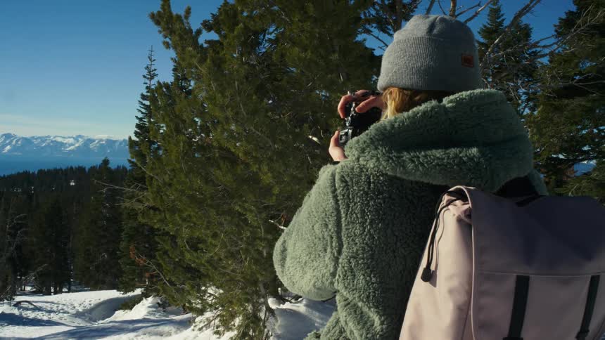 Woman with gray wooly hat green jacket and beige backpack photographs the snowy forest and mountain range in the snowy hill
