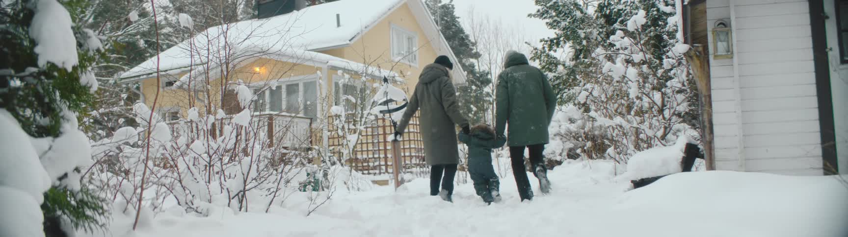 Father with green coat and black pants and mother in green overcoat hold toddler sons hands and walk through snow towards building