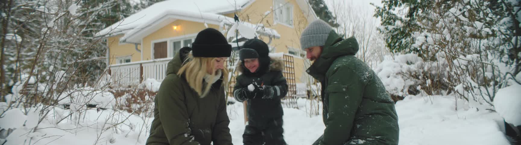 A mother and father gently throw snowballs at their young son and one another outside their home
