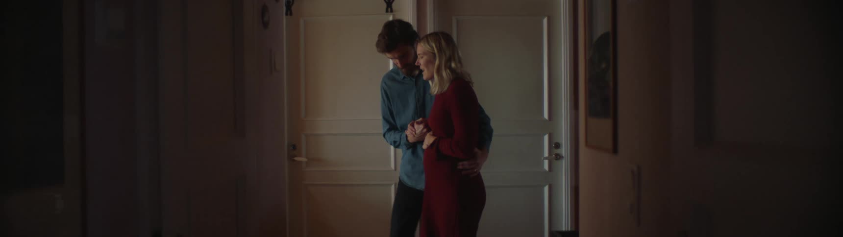 Man with shirt and beard helps blonde pregnant woman wearing red dress walk at home