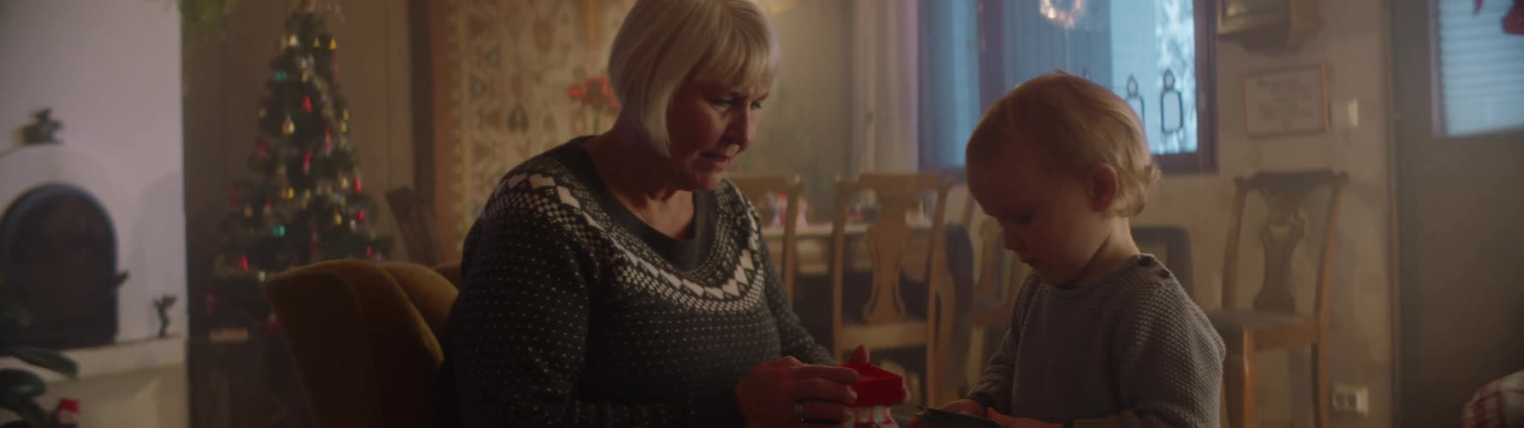 Grandmother with christmas jumper sitting in armchair plays with grandson with christmas tree in the background at home