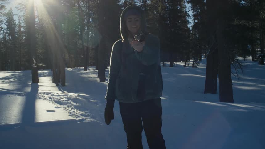 Woman wearing winter coat stands in snowy forest feeding birds by hand