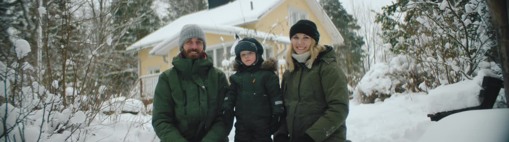 Young family wearing winter jackets posing in front of yellow cottage in the snow