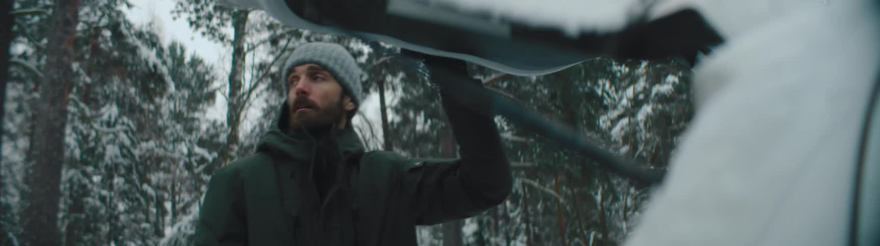 Man with gray wooly hat beard and green jacket takes bag from car in the snowy woods