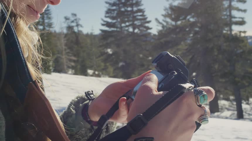 Blonde woman stands on snowy slope in conifer forest looking through pictures on digital camera