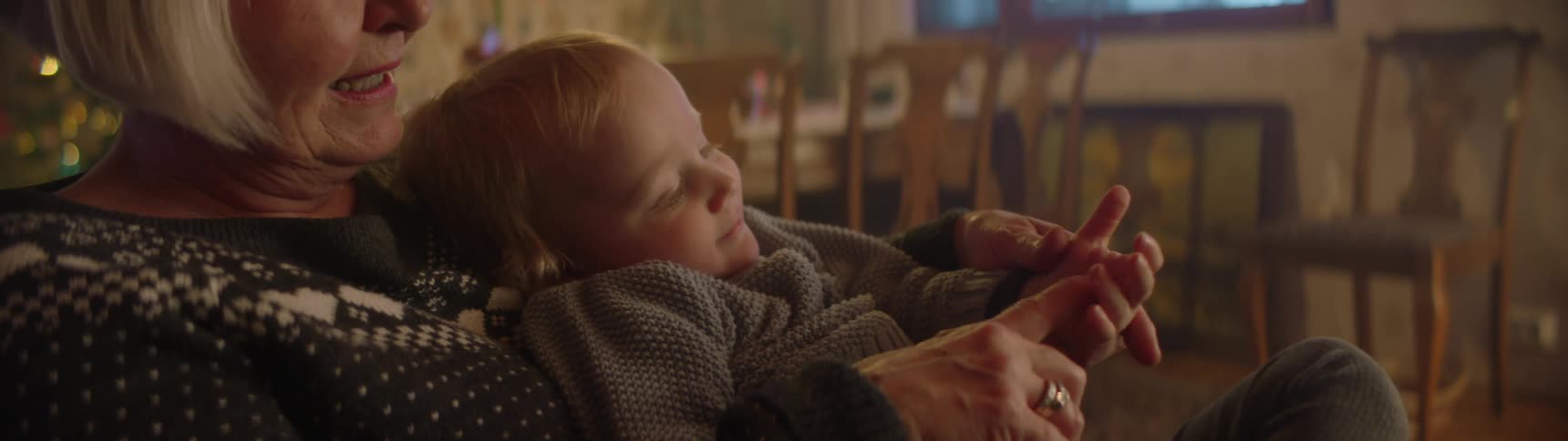 Grandmother with short fair hair and christmas sweater sits in armchair and holds grandson playing with him in living room at home