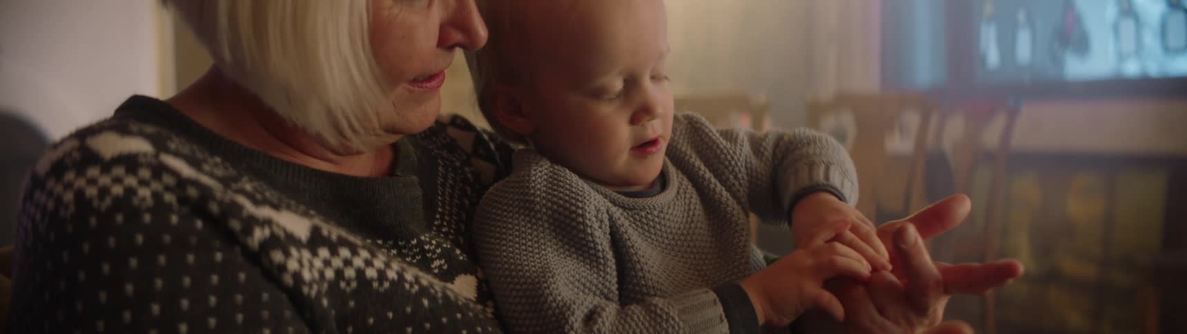 A woman at home holds her young grandson in her lap as he laughs and plays with her fingers