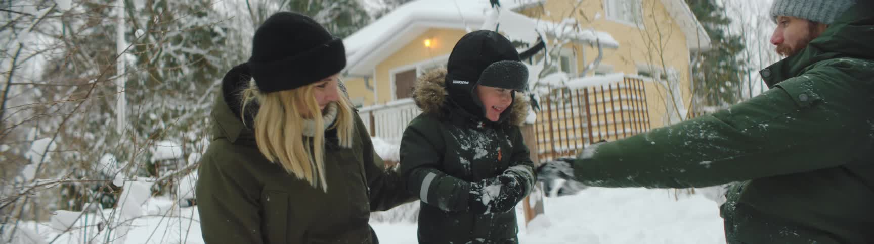 Mother father and child playing in the snow in front of a yellow cottage in the woods